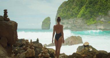 Siyah mayo giymiş bir kadın sahilde durup denize bakıyor. Tatilde rahatlayan ve deniz manzarası seyreden bir kız. Nusa Penida, Bali, Endonezya 'daki Tembeling Plajı ve Ormanı.