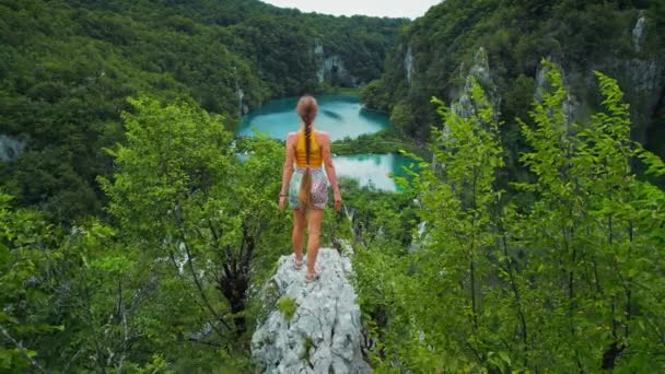 运动员攀岩少女在山顶上 望着碧绿的湖水和高高的瀑布 克罗地亚普里特维奇湖国家公园的女游客 夏天的冒险 — 图库视频影像