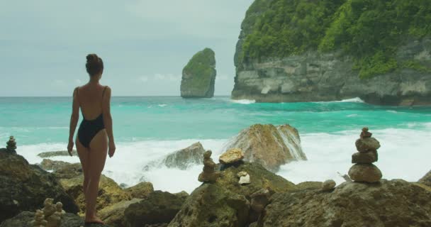巴厘岛Tembeling海滩上 一名身穿黑色泳衣的有反思能力的女人在岩石海岸上停下脚步 海浪在她身后颠簸着 Nusa Penidas悬崖 — 图库视频影像