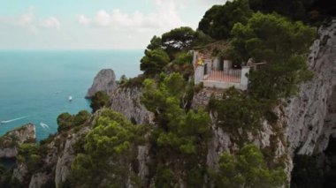 Bir insan yoğun çam ağaçlarıyla çevrili bir balkon kenarında oturur. Aşağıda, deniz ufka doğru uzanıyor, uçurumlar derin mavi sulara dökülüyor..
