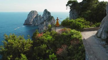 Capri Adası 'nın büyüleyici deniz manzarasına tepeden bakan sarı elbiseli kadın. Iconic Faraglioni deniz yığınları doğanın güzelliğiyle çevrili...