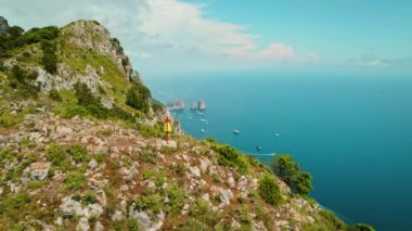 Capri Adası 'nın nefes kesici hava manzarası. Yeşil manzarada yürüyüş yapan bir kız. Denize ve dağınık teknelere bakıyor. Ünlü Faraglioni arka planda sallanıyor..