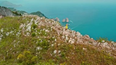 Yazlık elbiseli bir kadın, dağlarda yürüyüş yapıyor. Capri Adaları 'nın manzaralı güzelliğiyle, masmavi denize bakıyor. Arka planda ünlü üç uçurum Faraglioni...