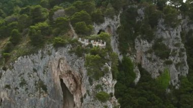 Uçurumun tepesinde denize bakan bir gözlem güvertesinde dururken turist kameralarına el sallıyor. Balkonu olan bir mağaranın tepesinde yemyeşil kayalar kulesi...