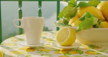 Açık bir masada kesilmiş bir limonun yanında el değmemiş bir fincan sıcak çay duruyor. Huzurlu sahne, canlı bir tekstil üzerinde limonların arasında bir çay setini gösteriyor...