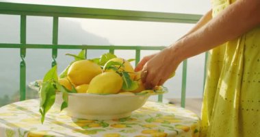 Dişi eller güneşli bir masada bir kase limon düzenler. Parlak, sıcak parıltı terasta taze narenciye ve yeşil yaprakları vurguluyor...
