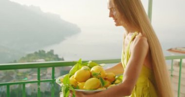 Okyanus manzaralı bir balkonda limon düzenleyen bir kadın. Dişi bir kase turunçgil meyvesini masaya yerleştiriyor, deniz zemininde parlıyor. Yaz İtalya..