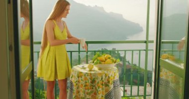 Balkonda içki içen bir kadın, masada limonlar, görünürde Amalfi sahilleri. Dişi, yanında narenciye olan bir içecek alır...