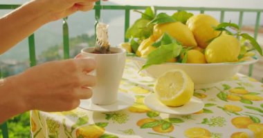 Çay poşeti beyaz bir kupada limonlarla dolu bir masada, dağların ötesinde. Daldırma hareketi güne huzurlu bir başlangıcı yansıtır. Kadın ellerine yakın çekim..