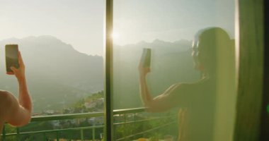 Kadın silueti cama yansıyor, akıllı telefon deniz manzarasını hedefliyor. Turist kız sabahın erken saatlerinde balkondan deniz manzarası çekiyor. İtalyan Tatilleri..