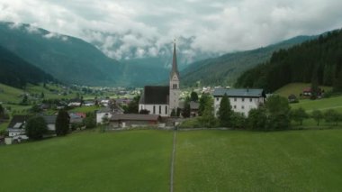 Avusturya 'nın Gosau köyü, ikonik kilise kulesi yeşil çayırlar ve kırsal binalar arasında yükseliyor. Sisle kaplanmış görkemli dağlar dramatik bir zemin oluşturur...