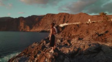 Bir çift kayalık arazide durur, güneşin okyanusun üzerinde batışını izlerken kucaklaşarak, parlayan gökyüzüne karşı bir siluet oluştururlar...