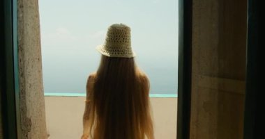 Uçsuz bucaksız mavi okyanusa bakan terastan dışarı bakan bir kadın. Uzun saçları, panoramik deniz manzarasının tadını çıkarırken akıyor...
