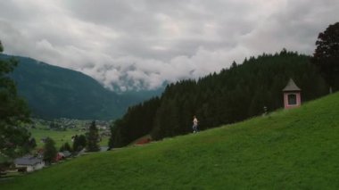 Avusturya, Gosau 'da yeşil bir tepe boyunca yürüyen bir yürüyüşçüyle sakin bir dağ manzarası. Arka planda yoğun ormanlar ve sis kaplı dağlar..