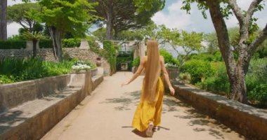Kadın, tarihi taş işçiliğiyle güneşli bir bahçe yolunda yürüyor. Zengin yeşillikler ve eski çiçek aksanlarıyla çevrili turistler huzur içinde yürür...