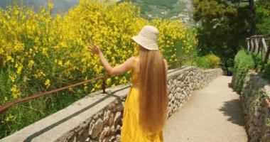 Yeşilliklerle çevrili, manzaralı bir patika boyunca canlı sarı çiçeklere dokunan bir kadın. İzler resim gibi bir manzaranın içinden geçiyor...