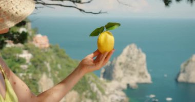 Kadın, Capri Sahili 'nin arka planında limon tutuyor. Faraglioni kayaları ve deniz mesafesi görünür.