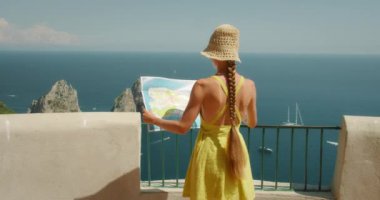 Elbise ve hasır şapkalı kadın güneşli bir günde İtalya 'nın Capri kentindeki Faraglioni kayalarına bakarken haritaya bakıyor...