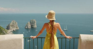 Hasır şapkalı ve sarı elbiseli bir kadın, İtalya, Capri Adası manzaralı bir manzaradan Faraglioni kayalarının ve derin mavi denizin manzarasına hayran.