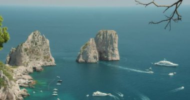 Capri 'deki Faraglioni kaya oluşumlarının açık mavi denizde güneşli bir gökyüzünün altında yüzen çeşitli tekne ve yatlarla görüntüsü...