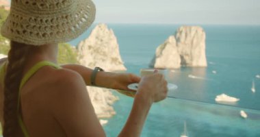 Kadın turist elinde deniz manzaralı bir kahve fincanı ve yaz günü Capri İtalya 'da Faraglioni uçurumları ile balkonda duruyor...