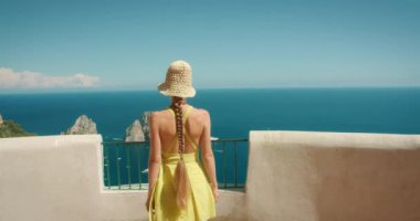 Sarı elbiseli ve şapkalı bir kadın. Uzakta Faraglioni kayaları ile Capri Denizi 'ne bakıyor.