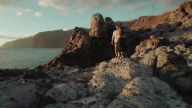 Bir kadın ve bir erkek Tenerife, İspanya 'daki Los Gigantes uçurumlarındaki kayalık sahil şeridinde durup arkalarında güneş batarken okyanusu seyrediyorlar...