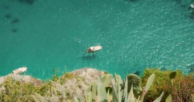 Açık havada yüzen bir tekne, kayalık bir uçurumun kenarında turkuaz bir su yemyeşil ve kaktüsü ile yaz güneşli bir günde ön planda...