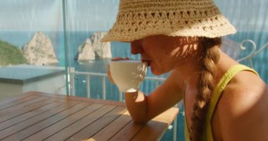 Hasır şapkalı kadın, güneşli bir günde Faraglioni kayalarına tepeden bakan ahşap bir masada otururken beyaz bardaktan kahve yudumluyor...