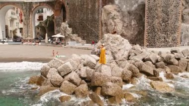 Kadın, Atrani kasabasındaki büyük kayalıklarda duruyor ve tarihi mimarisi arka planda görünüyor...