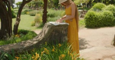 Sarı elbiseli bir kadın, yemyeşil bir bahçedeki taş bir yüzeye yayılmış bir harita üzerinde çalışıyor...