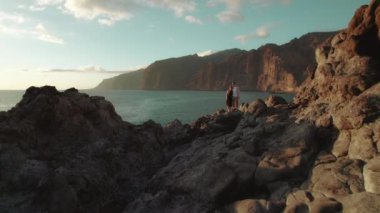 Tenerife 'deki Los Gigantes Kayalıkları' nda engebeli bir arazide duran bir çift, açık bir gökyüzü altında arkalarında dramatik bir şekilde yükselen volkanik kayalar...