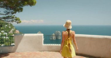 Capri Adası 'nın ve Faraglioni kayalarının çarpıcı sahil manzarasına hayran bir kadın elbisesi ve hasır şapkası giymiş...