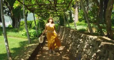 Neşeli bir kadın bahçedeki güneşli bir yaz gününde asma kaplı bir pergola altında yürüyor...