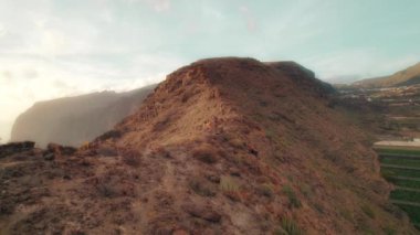 Alacakaranlıkta kayalık tepeler boyunca koşan kişi, engebeli uçurumlar ve okyanus manzarasıyla Tenerife, Kanarya Adaları, İspanya 'da puslu bir günbatımı gökyüzünün altında..