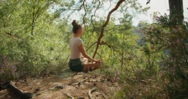 Bir kadın kayanın üzerinde bağdaş kurmuş, sakin bir ormanda nefes kesici bir uçurum manzarasıyla meditasyon yapıyor..