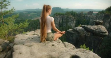 Kayalık bir çıkıntıda meditasyon yapan bir kadın. Uçurumların, ormanın ve mavi gökyüzünün altındaki uzak manzaranın panoramik manzarasına bakıyor.