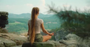 Kadın bir kayanın üzerinde oturuyor, sırtı kameraya dönük meditasyon yapıyor, Bastei 'nin geniş ve pitoresk manzarasına bakıyor...