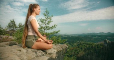 Meditasyon yapan bir kadının yan profili kayalık bir çıkıntıda duruşu, geniş bir orman manzarası ve yuvarlanan tepeler.