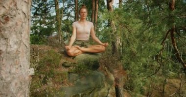 Kadın, sakin bir orman ortamında uzun ağaçlar ve yeşilliklerle çevrili bir kayanın üzerinde bağdaş kurarak meditasyon yapıyor..