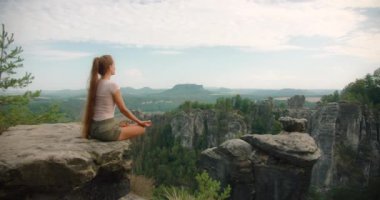 Bir kayanın üzerinde meditasyon yapan bir kadının geniş bir ormanı ve açık bir gökyüzünün altındaki kayalık araziyi seyredişinin yan görüntüsü.
