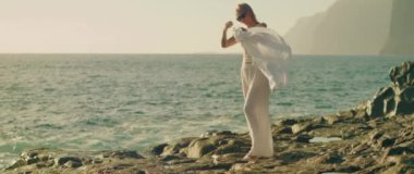 Okyanusun kıyısındaki kayalık bir sahilde dalgalı beyaz elbiseli bir kadın. Yüzüne hoş bir rüzgar esiyor..