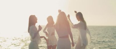 Bir grup kadın kumsalda şenlikli bir anın tadını çıkarıyorlar, saçları rüzgarda uçuşurken birlikte gülüyor ve dans ediyorlar...