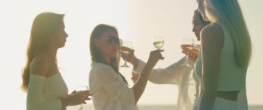 Kadınlar, okyanus kıyısında kutlama yaparken yüzleri güneş ışığıyla aydınlanarak hafif bir kadeh kaldırır. Dört arkadaş samimi bir anı paylaşıyor, kumsalda gün batımında şarapla kadeh kaldırıyorlar...