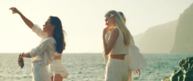 Beyaz elbiseli kadınlar, deniz kenarında kaygısız bir anın tadını çıkarıyorlar. Dalgalar arka planda, berrak bir gökyüzünün altında yavaşça çarpıyor.