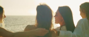 Kıyı şeridinde bir arada duran kadınlar, arka planda yumuşak okyanus dalgalarıyla güneşin tadını çıkarıyorlar. Dostlar kadeh kaldırır, günbatımının tadını çıkarırlar...