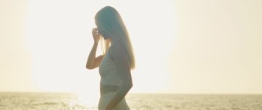 Deniz kenarında parlak güneş parıltılarıyla duran bir kadının arka plan manzarası, dış hatlarını ve sakin tavırlarını vurguluyor..