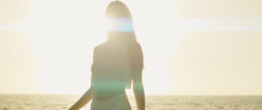 Güneş ışığı vücudunun etrafına yayılırken, deniz kenarına uzanan bir kadını sakin bir ifadeyle çerçeveler..