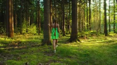 Yeşil elbiseli bir kadın, uzun ağaçlar ve canlı yeşilliklerle çevrili, güneşli bir ormana girer. Huzurun tadını çıkarır.