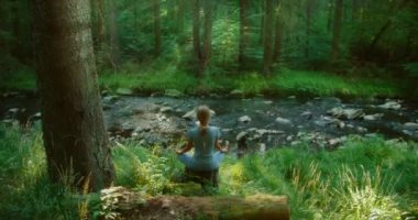 Kadın, orman akıntısına bakan bir ağaç kütüğü üzerinde meditasyon yapıyor. Etrafı yeşillik ve doğal sükunetle çevrili..